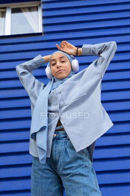 Baixo ângulo da mulher muçulmana em fones de ouvido modernos com a mão atrás da cabeça olhando para a câmera contra a parede do edifício urbano — Fotografia de Stock