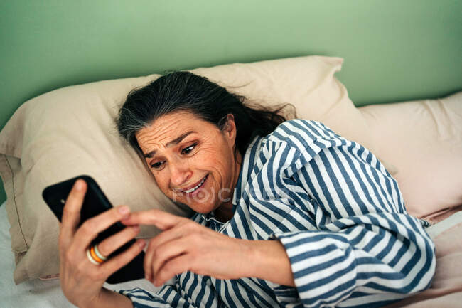 Von oben eine staunende hispanische Frau mittleren Alters mit langen dunklen Haaren in gestreifter Nachtwäsche, die glücklich lächelt, während sie gute Nachrichten auf dem Smartphone im Bett liegend liest — Stockfoto