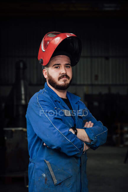 Серьезный бородатый рабочий в защитной каске и голубой в целом, стоя в гараже и глядя на камеру со скрещенными руками — стоковое фото