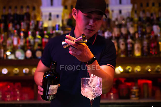 Junger asiatischer Barkeeper gießt Gin aus dem Jigger ins Glas, um in der Bar einen Gin Tonic Cocktail zuzubereiten — Stockfoto