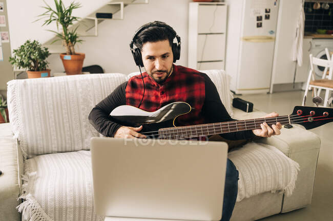 Musicien masculin adulte dans les écouteurs jouant de la guitare basse contre netbook sur canapé dans le salon — Photo de stock