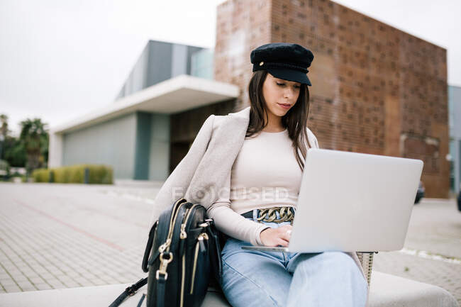 Concentrated young female entrepreneur in trendy outfit sitting on bench and browsing laptop while working on remote project in city — Stock Photo