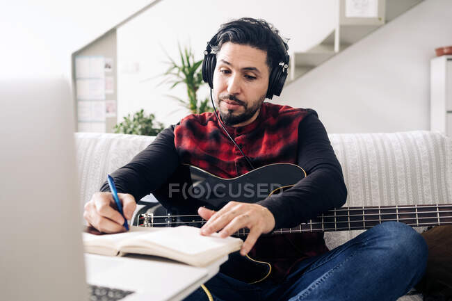 Guitarrista masculino adulto en auriculares con guitarra eléctrica tomando notas en cuaderno mientras compone música contra netbook en sofá en casa - foto de stock