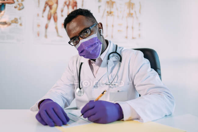 Médico afroamericano en anteojos trabajando mientras escribe en archivo de paciente en la mesa en el hospital - foto de stock