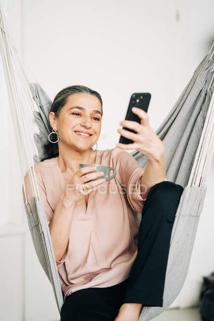 Позитивна жінка середнього віку з чашкою гарячого напою, що має відеочат на смартфоні, сидячи в зручному гамаку вдома на розмитому фоні — стокове фото