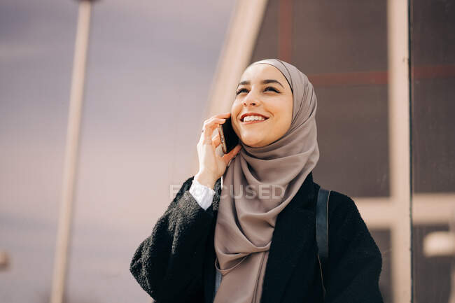 Позитивна етнічна жінка в хіджабі, що стоїть на міській вулиці і розмовляє на мобільному телефоні, дивлячись вгору — стокове фото