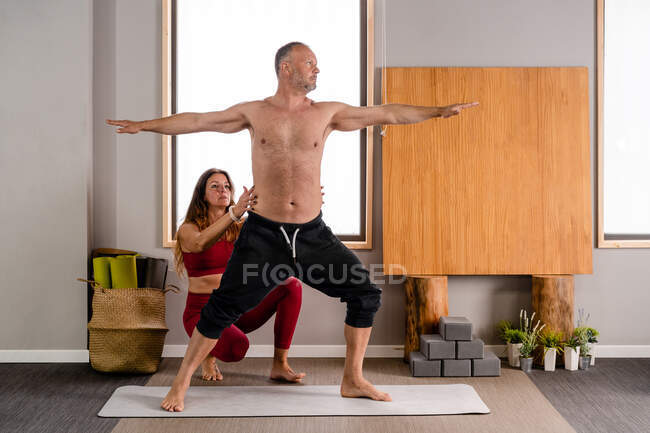 Entrenadora femenina concentrada en ropa deportiva enseñando a hombre realizando pose Virabhadrasana durante sesión de yoga en estudio - foto de stock