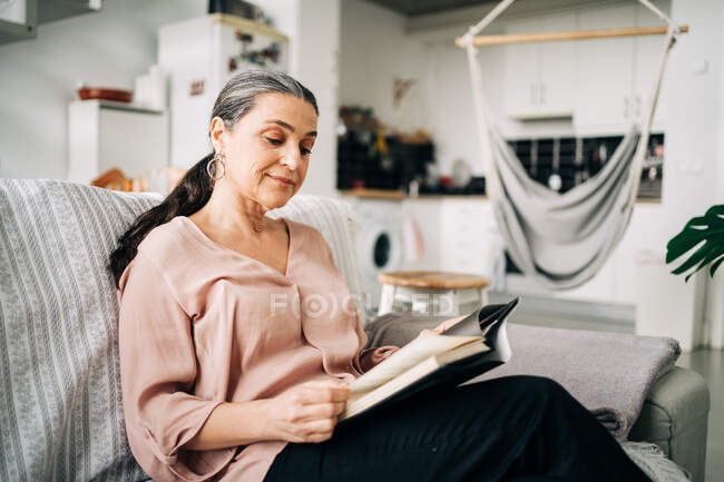 Livro de leitura feminina de meia idade concentrado enquanto sentado no sofá em apartamento moderno com balcão de cozinha e rede em casa — Fotografia de Stock