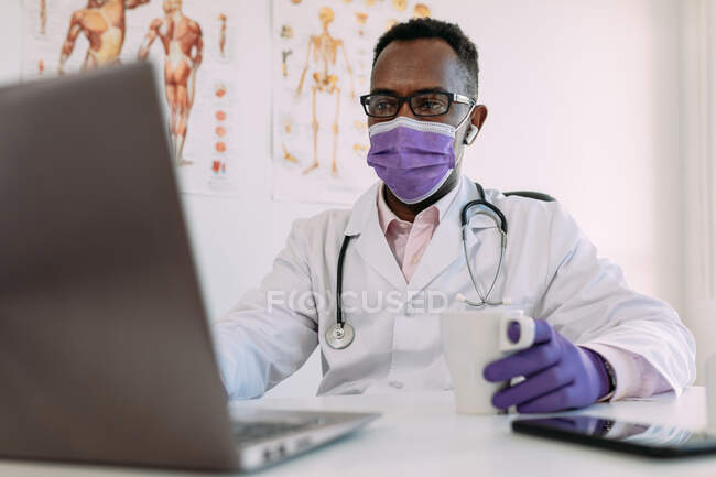 Médico masculino afroamericano concentrado irreconocible en bata médica y máscara beber café y trabajar en el ordenador portátil en la clínica moderna - foto de stock