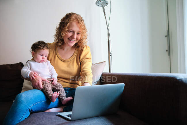 Веселая мать-одиночка фрилансер сидит с маленьким ребенком на диване и просматривает нетбук дома в дневное время — стоковое фото
