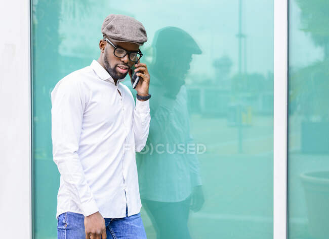 African American male in trendy outfit standing near glass building in city and talking on mobile phone — Stock Photo