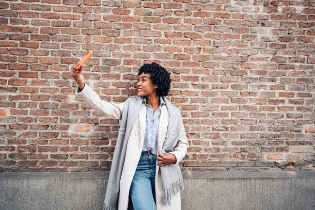 Позитивна афро-американська жінка в повсякденному одязі і пальто посміхається з радістю, роблячи автопортрет на мобільному телефоні. — стокове фото