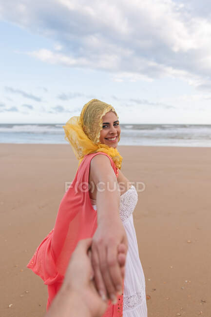 Fröhliche junge Frau lächelt und streckt anonymen Freund die Hand aus, während sie am sandigen Ufer des Ozeans in die Kamera blickt — Stockfoto
