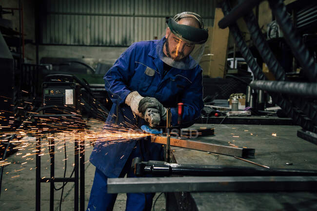 Arbeiter in Schutzmaske und blauer Arbeitskleidung schleift Metallteil mit Winkelschleifer an Werkbank in professioneller Garage — Stockfoto