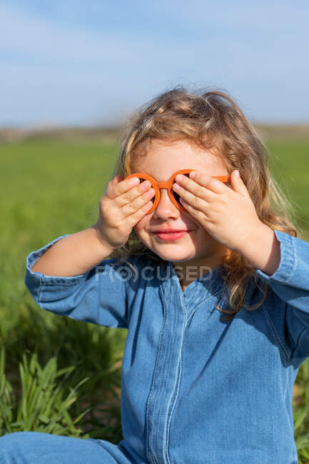 Adorable niña cubriendo los ojos mientras se sienta en el campo verde en verano y jugar al escondite juego - foto de stock