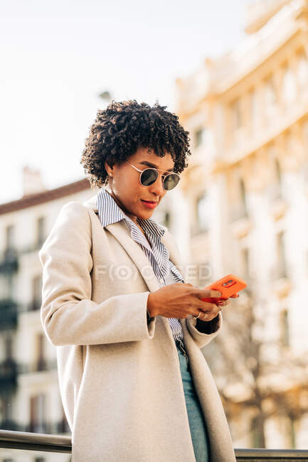 De baixo da mensagem de texto feminina afro-americana elegante no telefone celular enquanto caminhava na rua da cidade — Fotografia de Stock