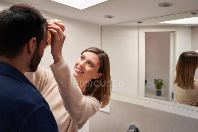 Feliz joven suéter casual femenino ajustando el pelo de novio barbudo irreconocible mientras está de pie cerca del espejo en el baño moderno - foto de stock