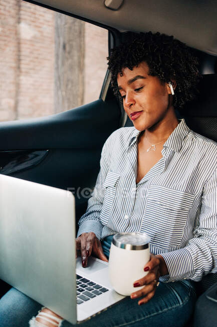 Junge, zufriedene Afroamerikanerin arbeitet mit Netbook und trinkt eine Tasse Kaffee, um auf dem Rücksitz im Auto zu fahren — Stockfoto