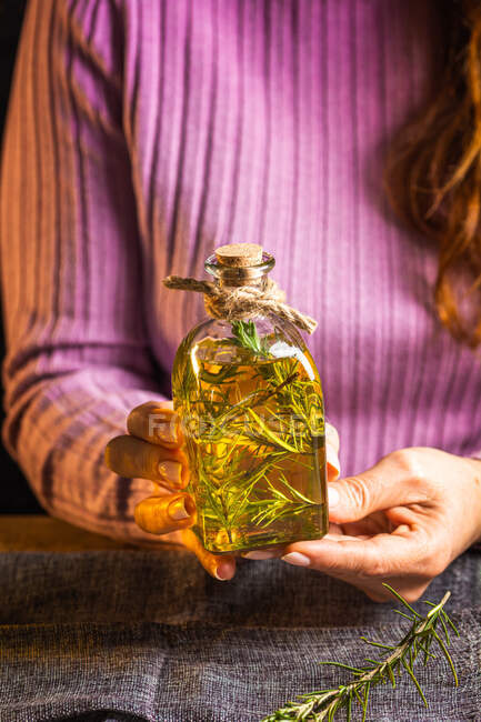 Cultiver dame anonyme en pull violet montrant bouteilles en verre à l'huile essentielle avec des brins d'herbes avec des feuilles vertes près du tissu sur la table — Photo de stock