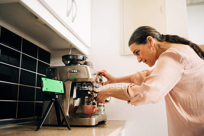 Вид збоку концентрованої жінки середнього віку, яка готує свіжу каву, використовуючи сучасну кавоварку на кухонній стійці зі смартфоном на тринозі — стокове фото
