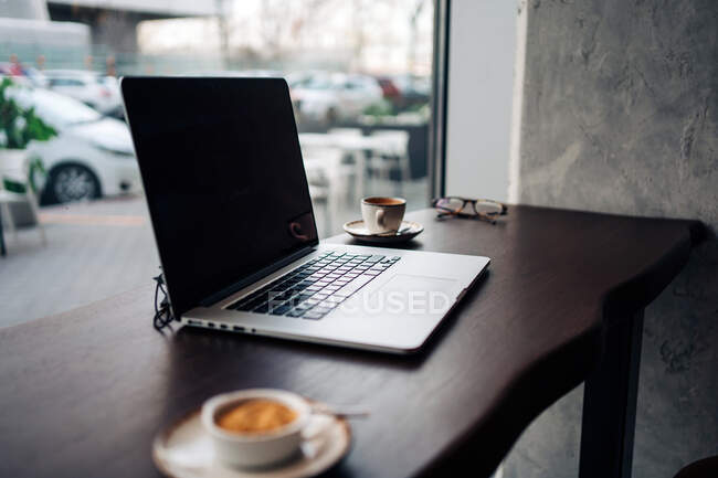 Netbook moderno con schermo nero su tavolo di legno con tazze di caffè aromatico in caffetteria — Foto stock