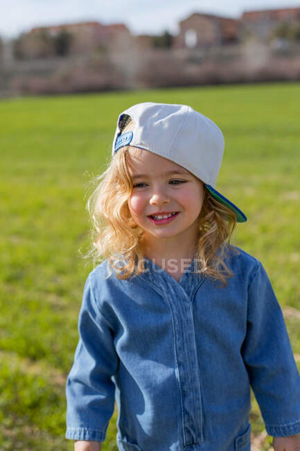 Glückliches Mädchen in stylischer Kleidung und Mütze schaut weg, während es an einem sonnigen Sommertag auf dem Feld auf dem Gras steht — Stockfoto