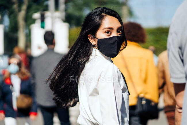 Back view of Asian female with flying hair wearing protective mask walking in crowd and looking at camera — Stock Photo