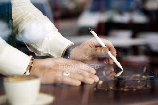 Through glass wall view of crop anonymous male executive using stylus and cellphone at cafeteria table — Stock Photo