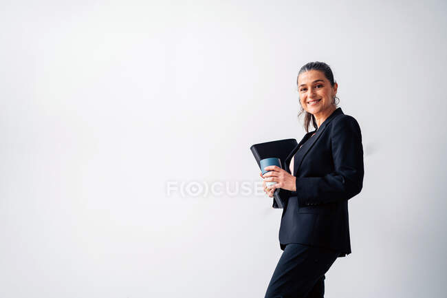 Positive middle aged female entrepreneur with ponytail wearing formal suit looking at camera while standing with folder and cup of drink on white background — Stock Photo