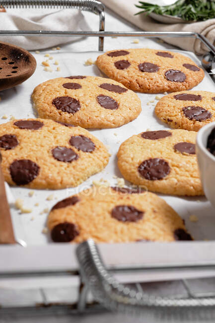 Desde arriba de galletas dulces recién horneadas con chips de chocolate en rejilla de metal colocados en la mesa con varias herramientas de cocina y ramas de romero verde - foto de stock