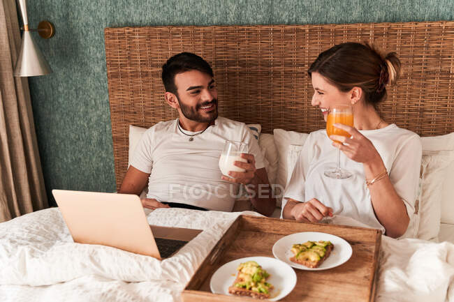 Счастливая молодая многонациональная пара в повседневной одежде завтракает и смотрит кино на ноутбуке, сидя в удобной постели во время ленивых выходных дома — стоковое фото