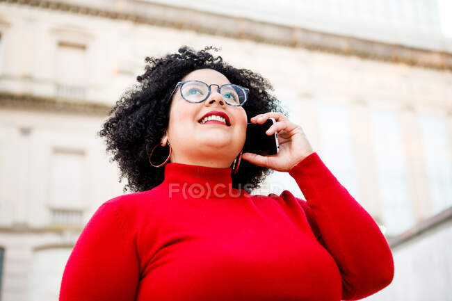 Знизу вмісту надмірної ваги жінка в червоному одязі та окулярах розмовляє на мобільному телефоні, дивлячись вгору по місту — стокове фото
