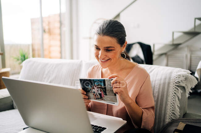 Femme assise sur un canapé et démontrant une photo tout en ayant un chat vidéo via un ordinateur portable dans le salon à la maison — Photo de stock