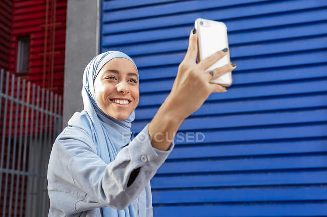 Femme ethnique amicale en foulard prenant autoportrait sur téléphone portable dans la rue de la ville au soleil — Photo de stock