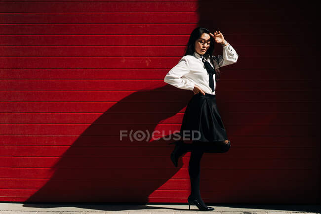 Full body side view of female with hand on waist covering face from sunshine standing against red gates — Stock Photo