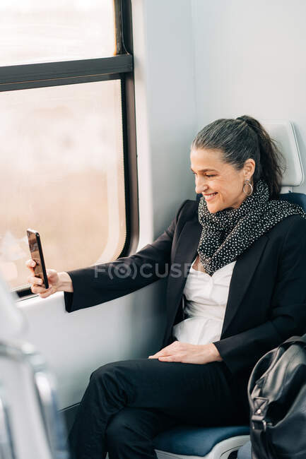 Side view of positive middle aged female with scarf taking picture on cellphone while sitting on passenger seat near window in wagon during journey — Stock Photo