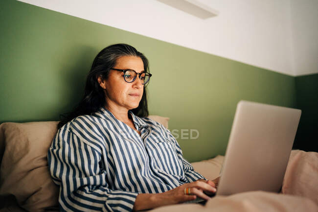 Freelancer étnica de mediana edad con pelo largo y oscuro en ropa casual y anteojos sentados en la cama y trabajando remotamente en el portátil en casa - foto de stock