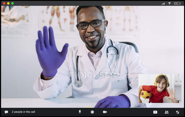 Niño feliz en los auriculares con la pelota en la mano y el joven afroamericano médico masculino saludándose con gesto de la mano, mientras que tienen video chat durante la consulta en línea - foto de stock