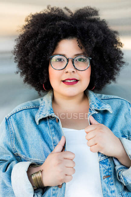 Adulto mulher com sobrepeso em roupas da moda e óculos com penteado afro olhando para a câmera no fundo borrado — Fotografia de Stock