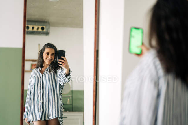 Mulher alegre de meia-idade vestindo blusa listrada refletindo no espelho ao tirar auto-retrato no smartphone no quarto em casa — Fotografia de Stock