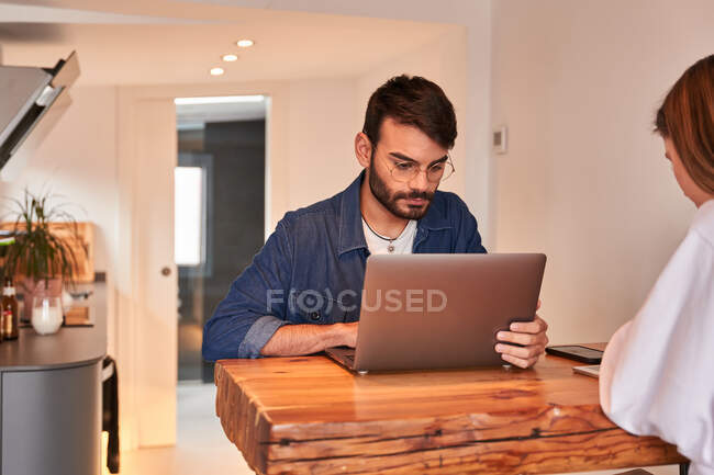 Freelancer masculino hispânico focado jovem trabalhando remotamente em laptop sentado à mesa na cozinha com namorada navegando smartphone — Fotografia de Stock