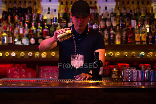 Joven camarero asiático vertiendo agua tónica al vaso para preparar un cóctel gin tonic en el bar - foto de stock