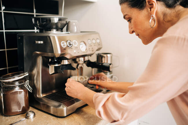 Side view of concentrated middle aged female preparing fresh coffee while using modern coffee maker at kitchen counter — Stock Photo