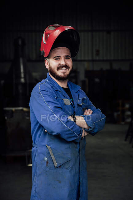 Happy bearded workman wearing protective hardhat and blue overall while standing in garage and looking at camera with crossed arms — Stock Photo