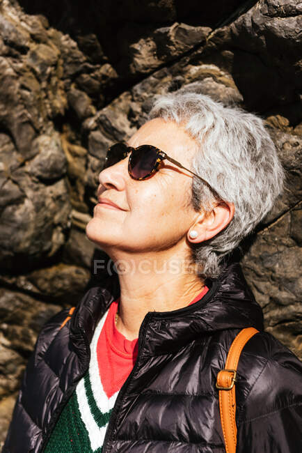 Вид збоку щасливої літньої жінки-туристки в повсякденному одязі, що стоїть на кам'янистому формуванні — стокове фото