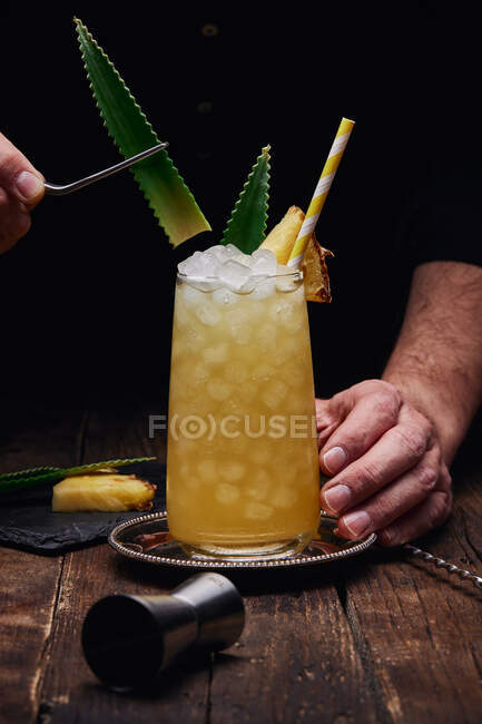 Crop anonymous barman garnishing alcohol cocktail with green leaves and pineapple piece on tray near shot glass at wooden table on black background — Stock Photo