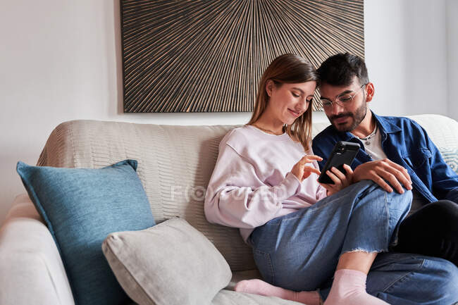 Romantic young diverse couple in casual clothes relaxing on sofa and watching photos on smartphone during weekend at home — Stock Photo