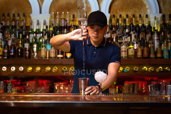 Jeune barman asiatique versant de la vodka dans le shaker tout en préparant un cocktail au bar — Photo de stock
