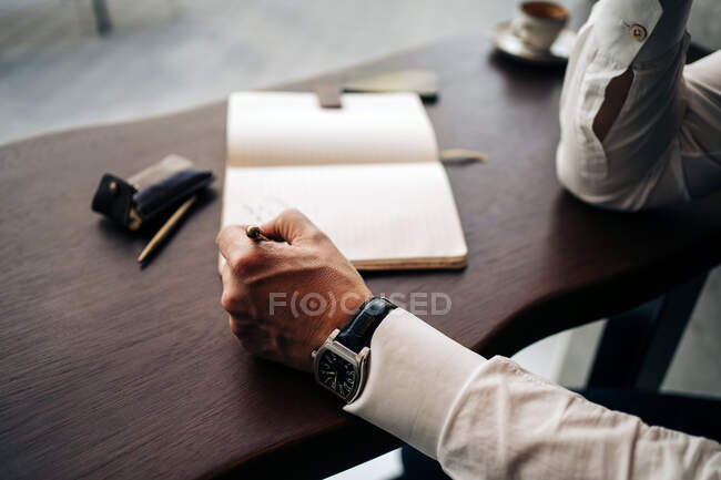 Crop anonymous businessman in wristwatch with pen and open journal working at cafeteria table in daylight — Stock Photo