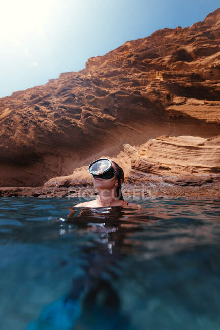 Female traveler in mask swimming in clean blue water against rocky cliff during trip — Stock Photo